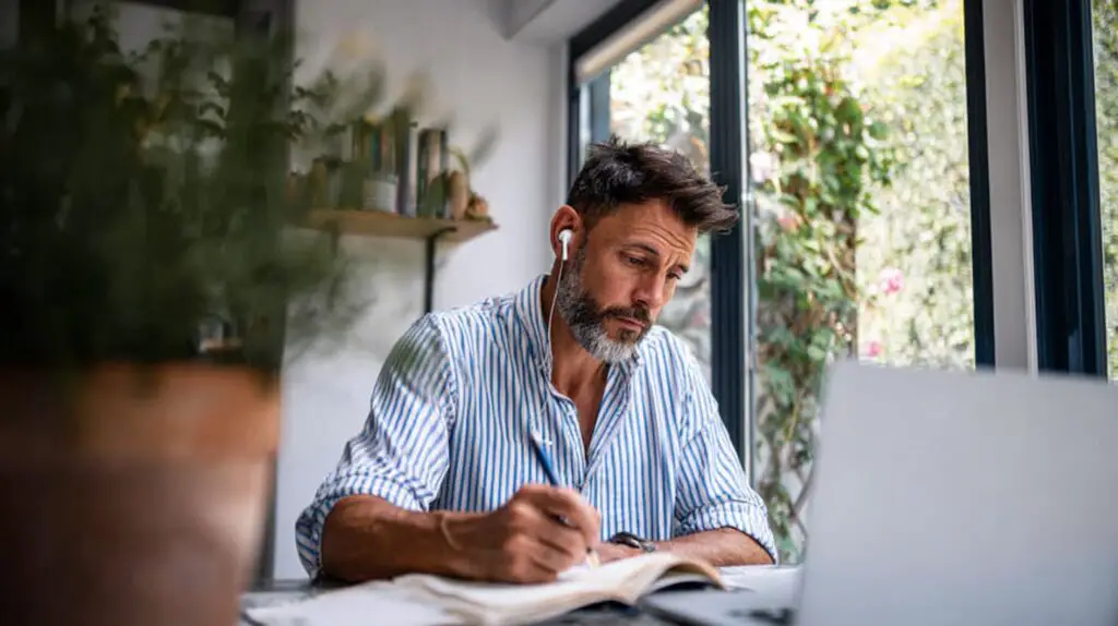  Freelance writer focused on laptop, jotting notes in a notebook; working in a bright home office.